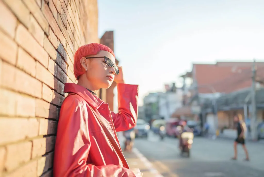 Google Search Engine Alternatives: A member of GenZ with short pink hair, classes and a red jacket, leaning up against a building on a sunny street