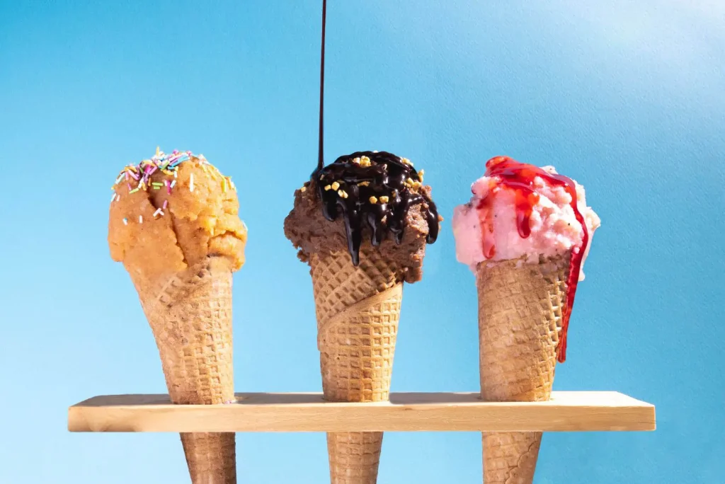 A small flight of three ice cream cones. On the left is a butter pecan flavor. Chocolate is in the middle with hot fudge drizzled on top. Strawberry is on the right.