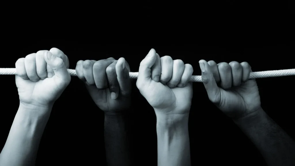 Four hands of varying skin tones grip a single white rope against a black background, symbolizing unity and cooperation.