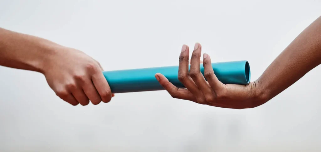 Close-up of one person handing a blue relay baton to another person against a plain background.