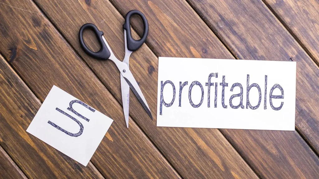 A pair of scissors cuts the prefix "un" from the word "unprofitable," leaving "profitable" on a wooden surface—a clever take on businesses that are growing but not profitable.